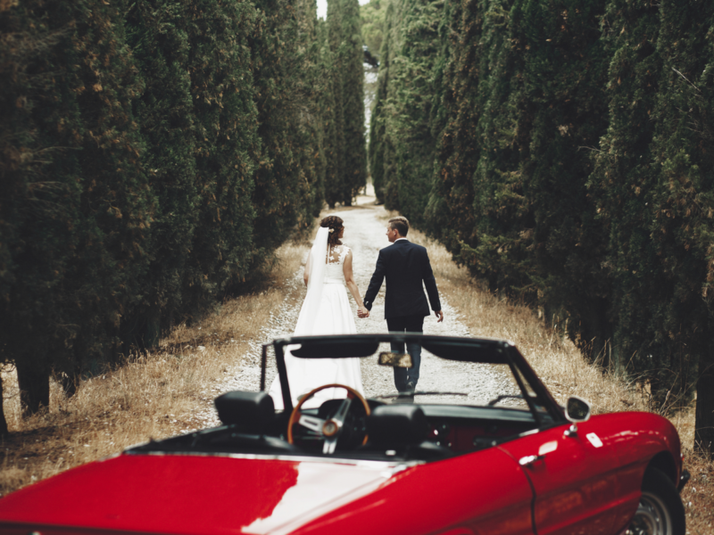 Bride and groom hold each other hands walking before a red cabrio between tall trees somewhere in Tuscany, Italy