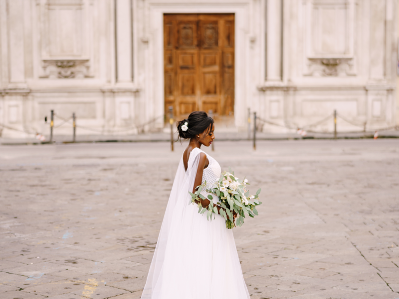 Interracial wedding couple. Wedding in Florence, Italy. African-American bride in a white dress with a bouquet in her hands, with a long veil stands on the street, against the background of the church