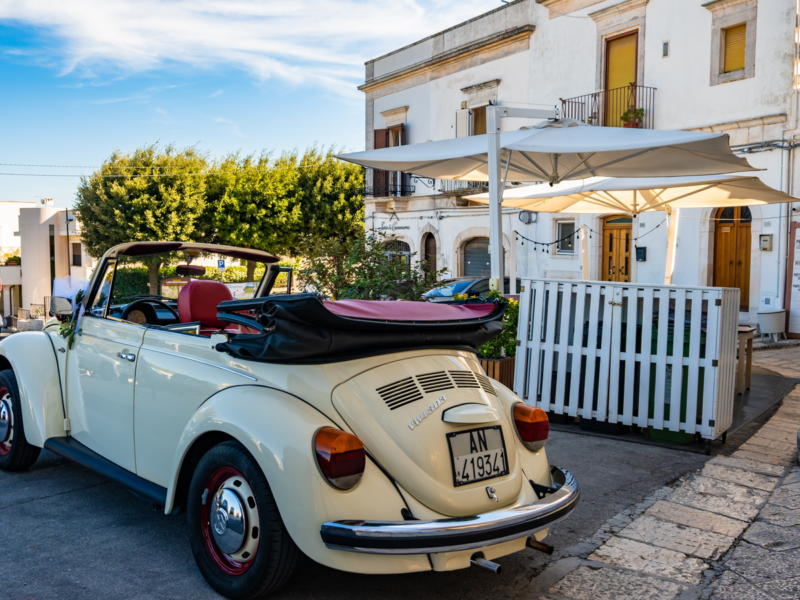 Vintage creme conversível carro Volkswagen Garbus estacionado perto de um café ensolarado com guarda-chuvas brancos prontos para a viagem de casamento, Apúlia, Itália